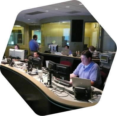A number of employees work at stations in the NOC while others confer in the background.